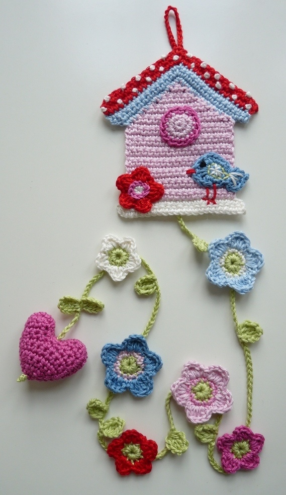 crocheted birdhouse with flowers and hearts hanging from it's side on a white surface