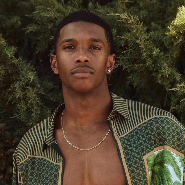 a man with no shirt standing in front of some trees and bushes wearing a necklace