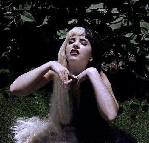 a woman with long blonde hair laying on the ground in front of some bushes and trees