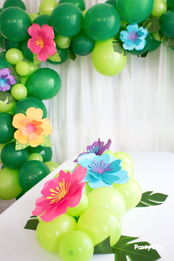 balloons and flowers are on the table in front of a balloon arch that is decorated with greenery