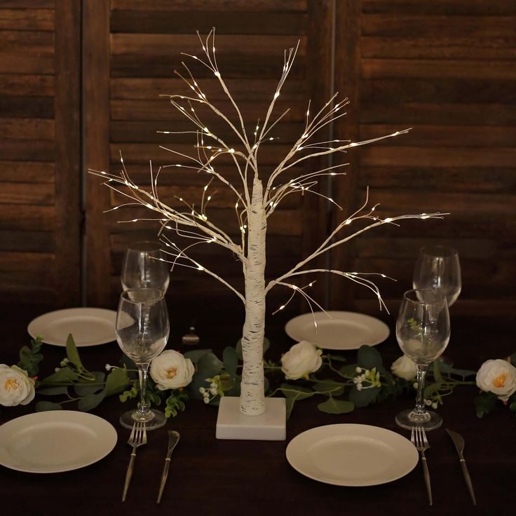 the table is set with white plates, silverware and a lighted tree centerpiece