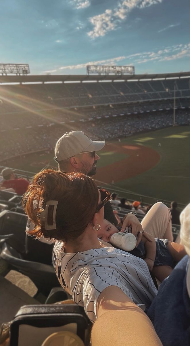 two people sitting in the stands at a baseball game, one is holding a baby