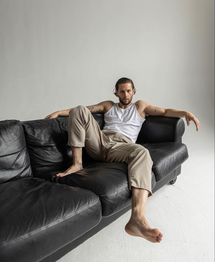 a man sitting on top of a black couch in front of a white wall with his legs crossed