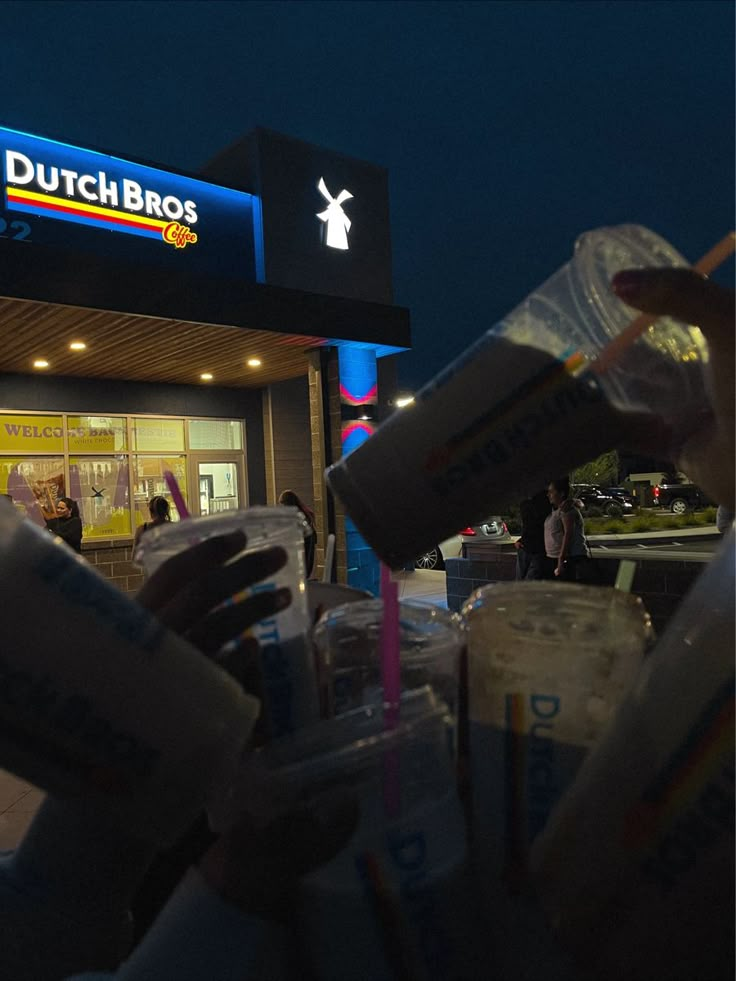 a person holding two cups in front of a dutch bros store at night with people walking by