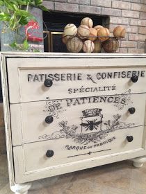 a white dresser with an antique french label on the front and bottom drawers, in front of a brick fireplace