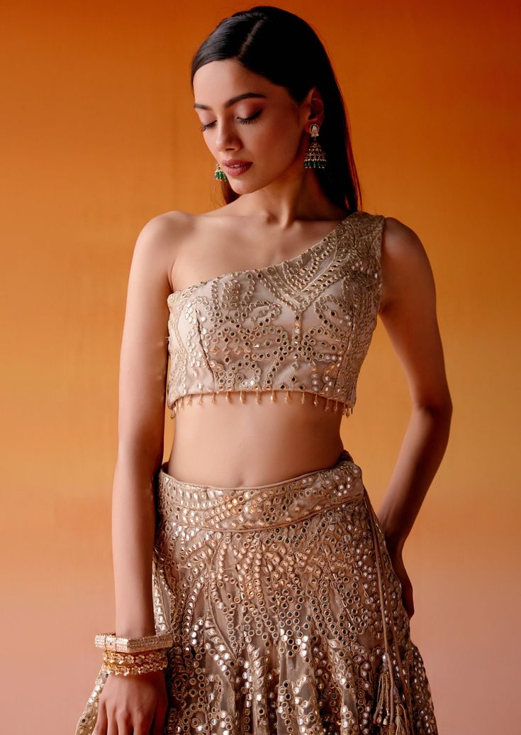 The lehenga is adorned with intricate mirrorwork and golden embellishments paired with an one-shoulder blouse in asymmetrical design, completed with mirrorwork detailing. One Shoulder Sequin Evening Sets, Embellished One-shoulder Evening Saree, Evening One Shoulder Embellished Pre-draped Saree, Evening One-shoulder Embellished Pre-draped Saree, One Shoulder Sequin Set For Party, One-shoulder Sequin Set For Party, One Shoulder Dress With Zari Work For Designer Wear, Traditional One-shoulder Set With Dupatta, Bollywood Style One-shoulder Fitted Pre-draped Saree