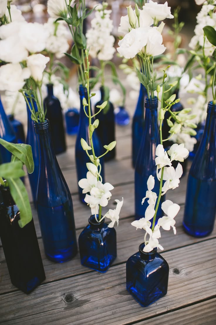 several blue vases with white flowers in them on a wooden table top, all lined up and ready to be used as centerpieces