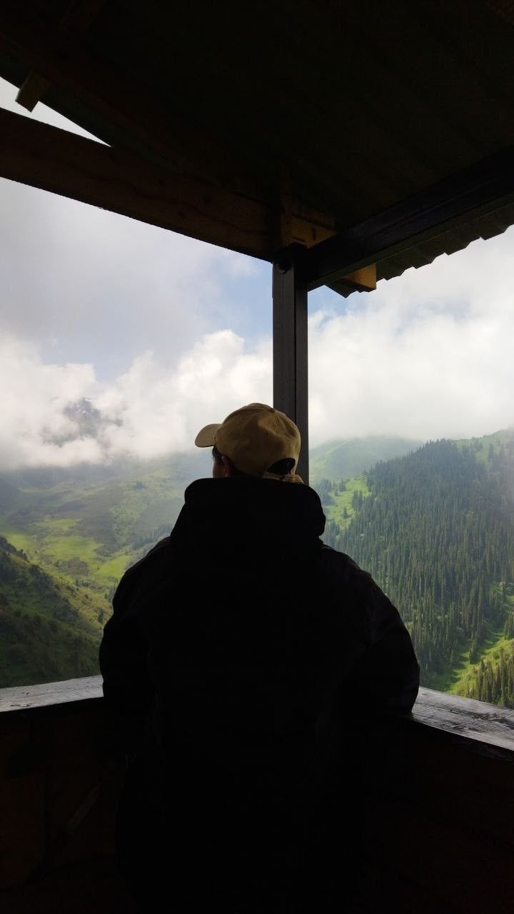 a man looking out the window at mountains and trees in the distance, while wearing a hat