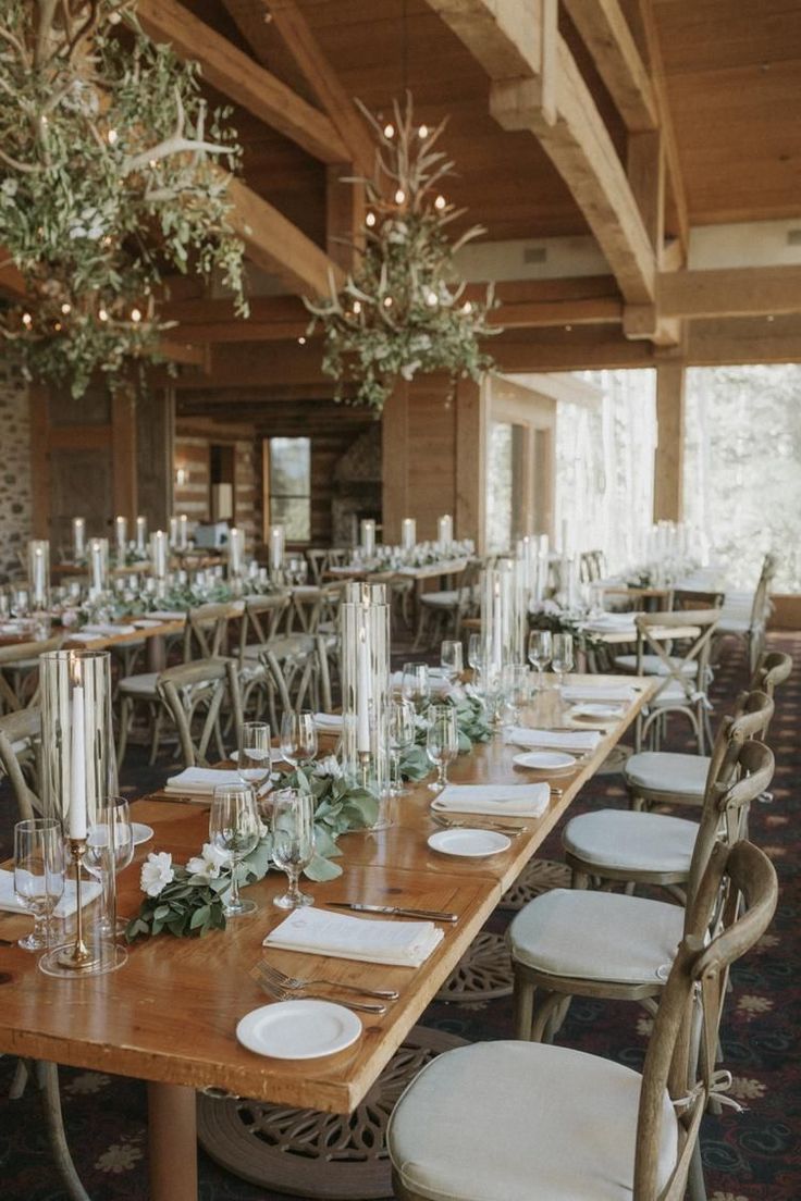 a long wooden table with white plates and silverware on it, surrounded by greenery hanging from the ceiling