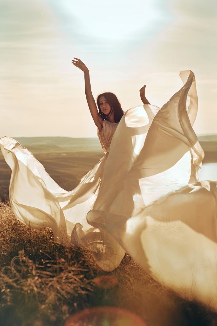 a woman in a white dress is standing on a hill with her arms spread out