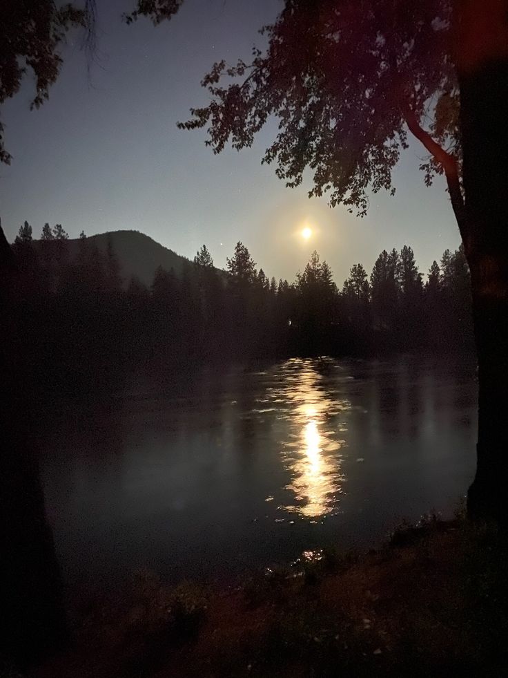 the moon is setting over a lake with trees in the foreground and mountains in the background