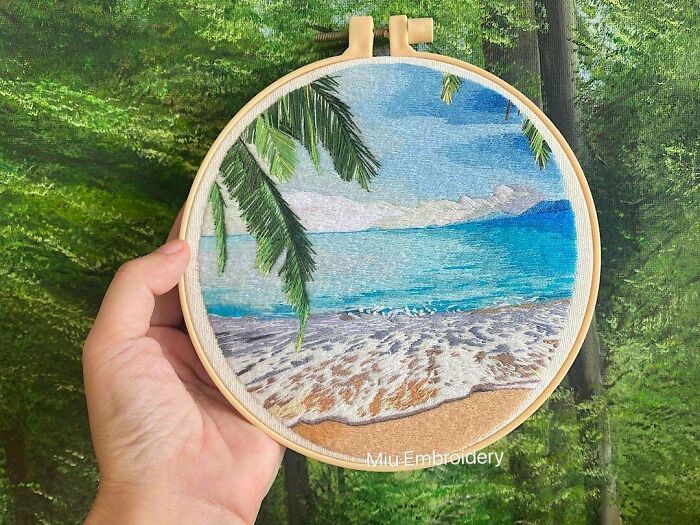 a person holding up a small embroidery art piece in front of some trees and the ocean