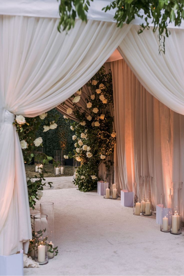 an outdoor ceremony with candles and flowers on the aisle, draped in white drapes