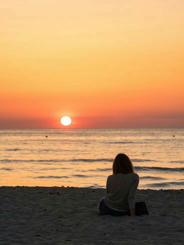 a woman sitting on top of a sandy beach next to the ocean under a sunset