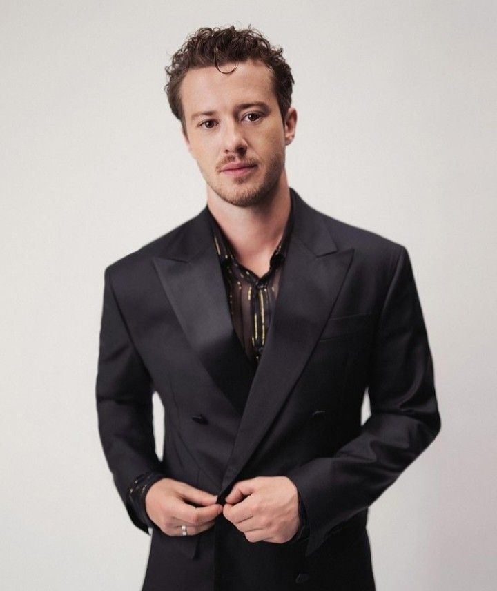 a man in a black suit and tie posing for a photo with his hands in his pockets