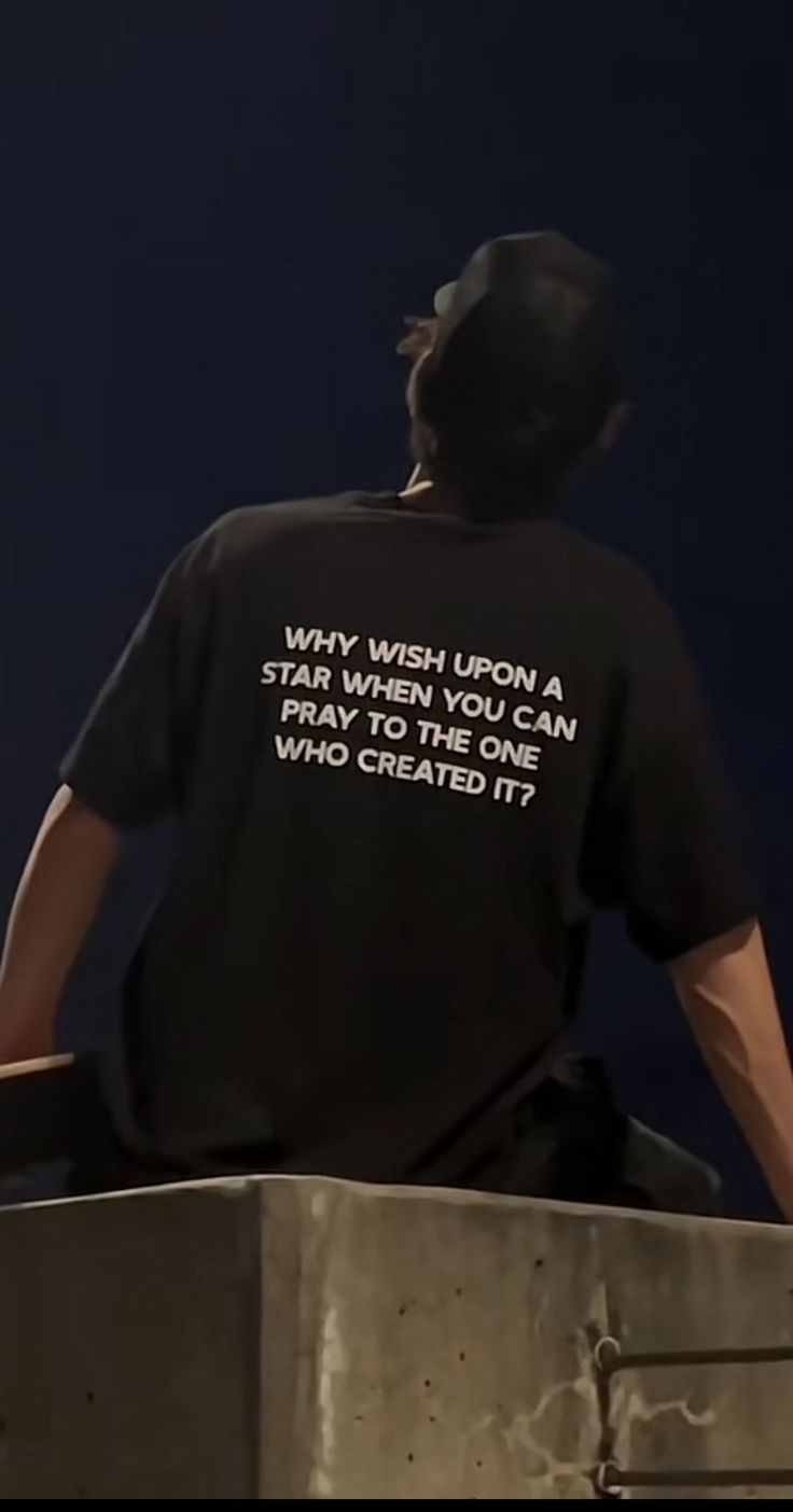 a man sitting on top of a cement block with his back to the camera, wearing a black t - shirt that says way wish upon a star you can pray to create who created it