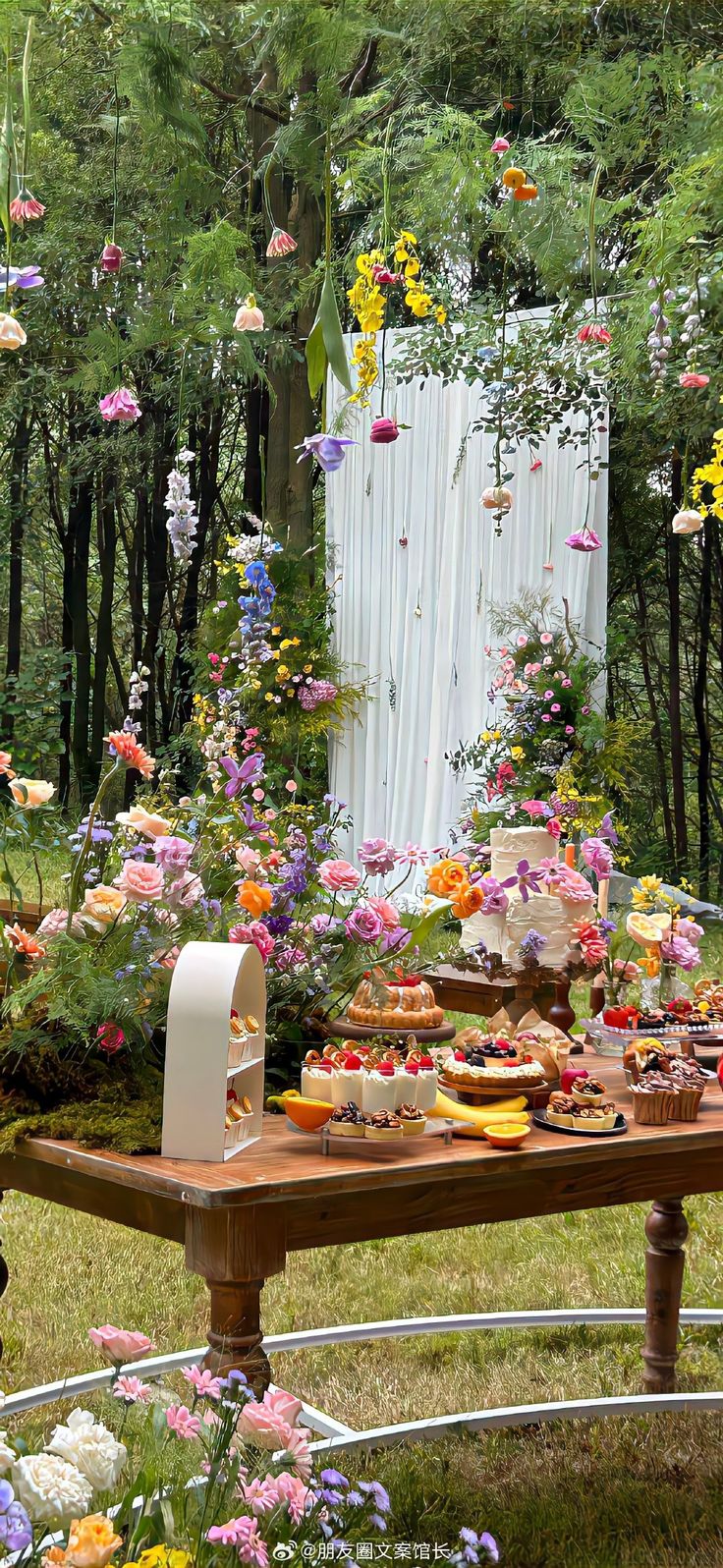 a table with flowers and cakes on it in the middle of a garden filled with trees