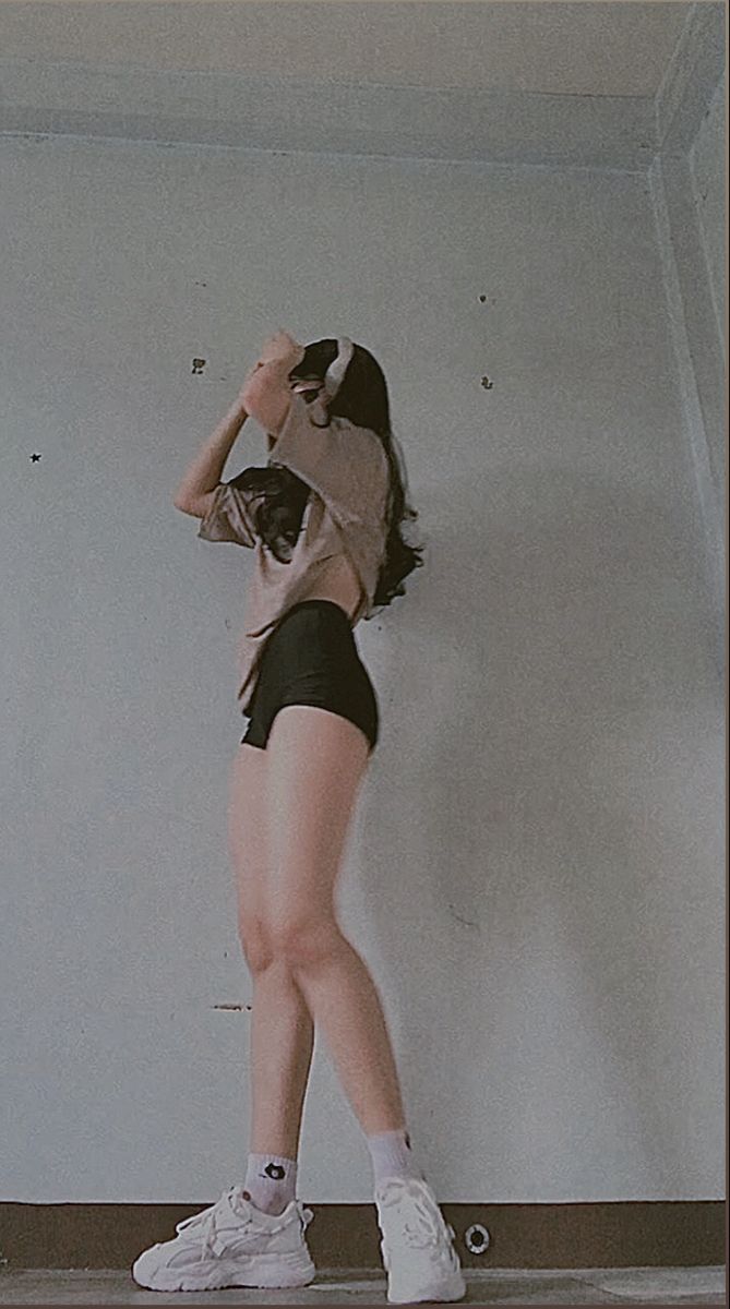 a woman standing on top of a wooden floor next to a white wall and holding her hands behind her head