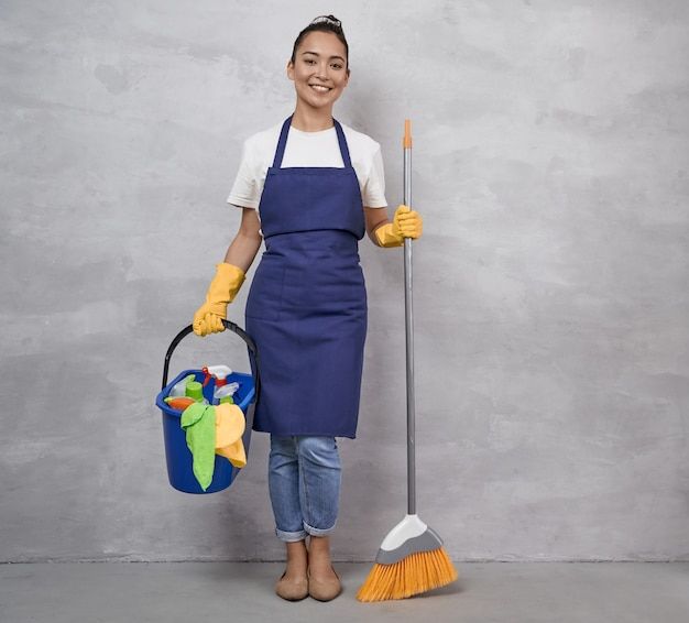 a woman in an apron and rubber gloves holding a mop, bucket and broom