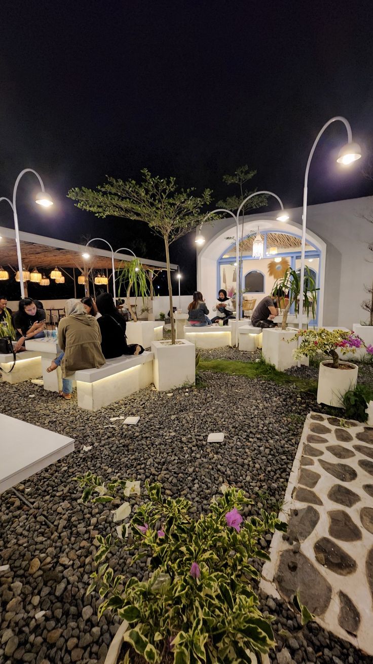 people are sitting on benches in the middle of a garden at night, with lights above them