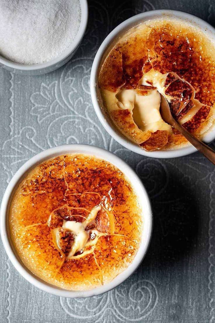 two white bowls filled with food on top of a gray tablecloth next to spoons