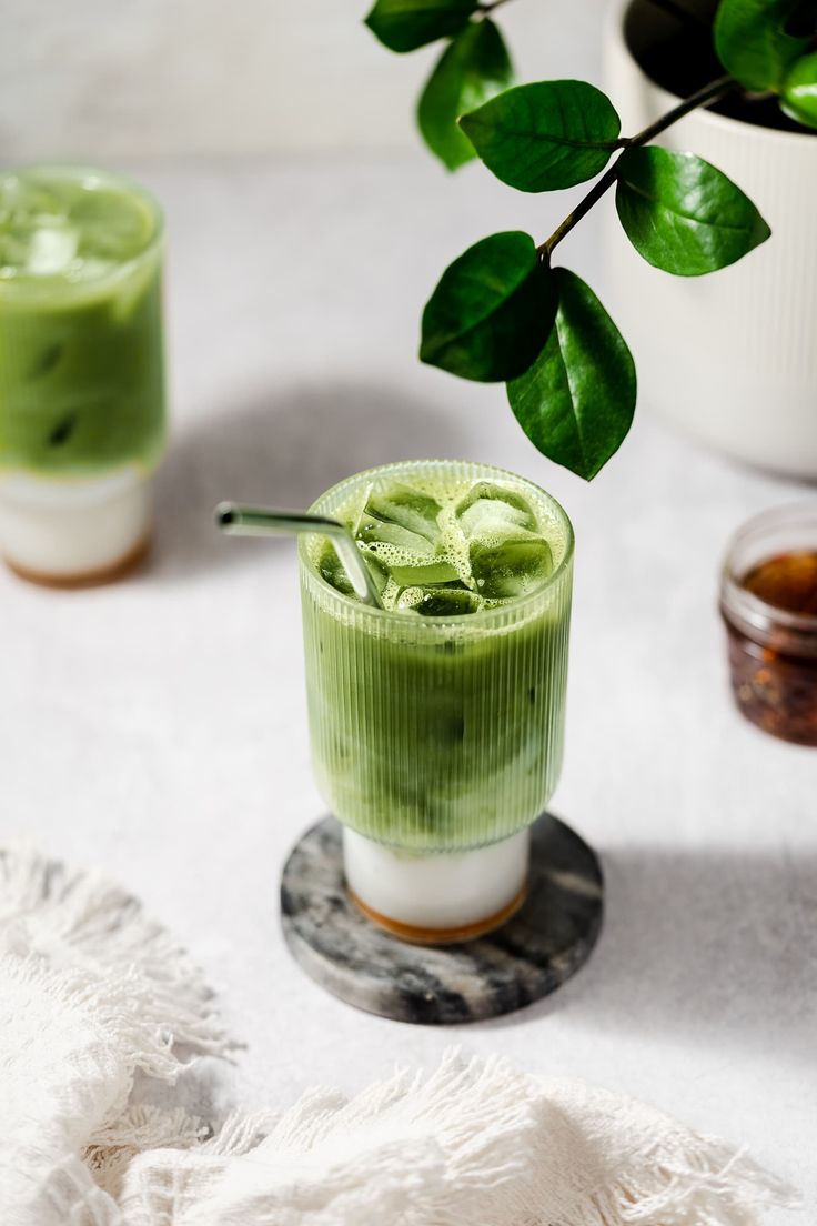 two glasses filled with green liquid next to a potted plant on a white table