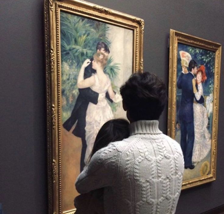 a man and woman looking at two paintings on display in an art gallery, one is wearing a white sweater