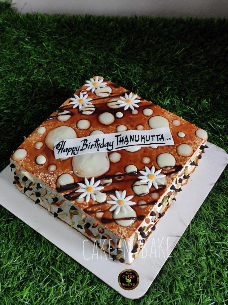 a square cake with white flowers on top sitting in the grass, and happy birthday thanuutta written on it