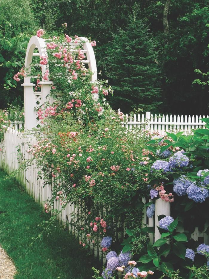 a white picket fence with pink and blue flowers growing on it next to a lush green garden