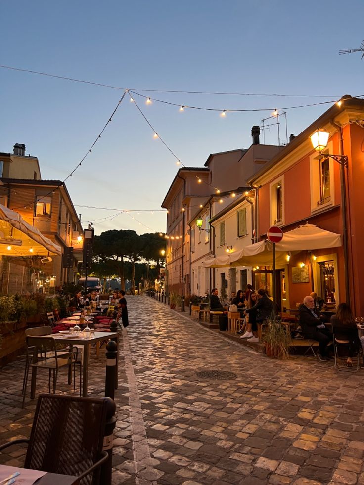 people are sitting at tables in an alleyway with string lights strung across the street