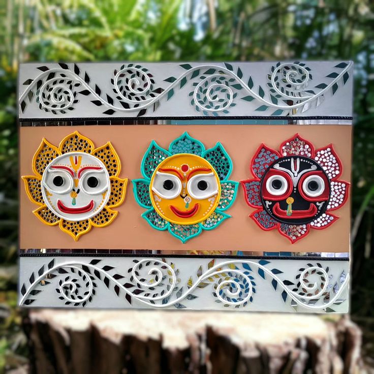 three colorful masks are sitting on top of a piece of wood in front of some trees