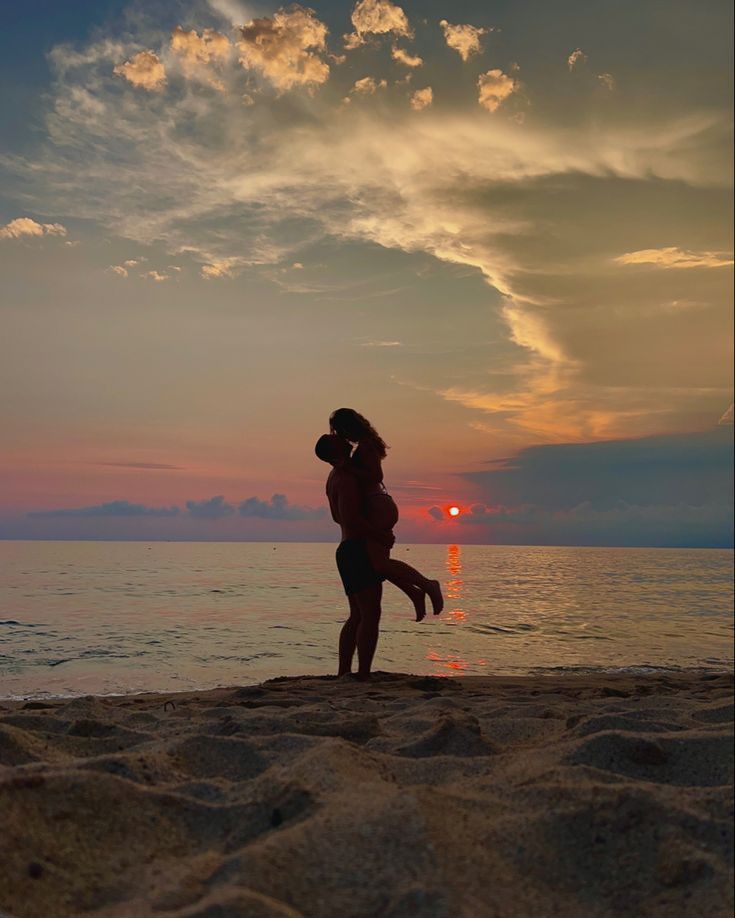 a man and woman kissing on the beach at sunset with clouds in the sky above them