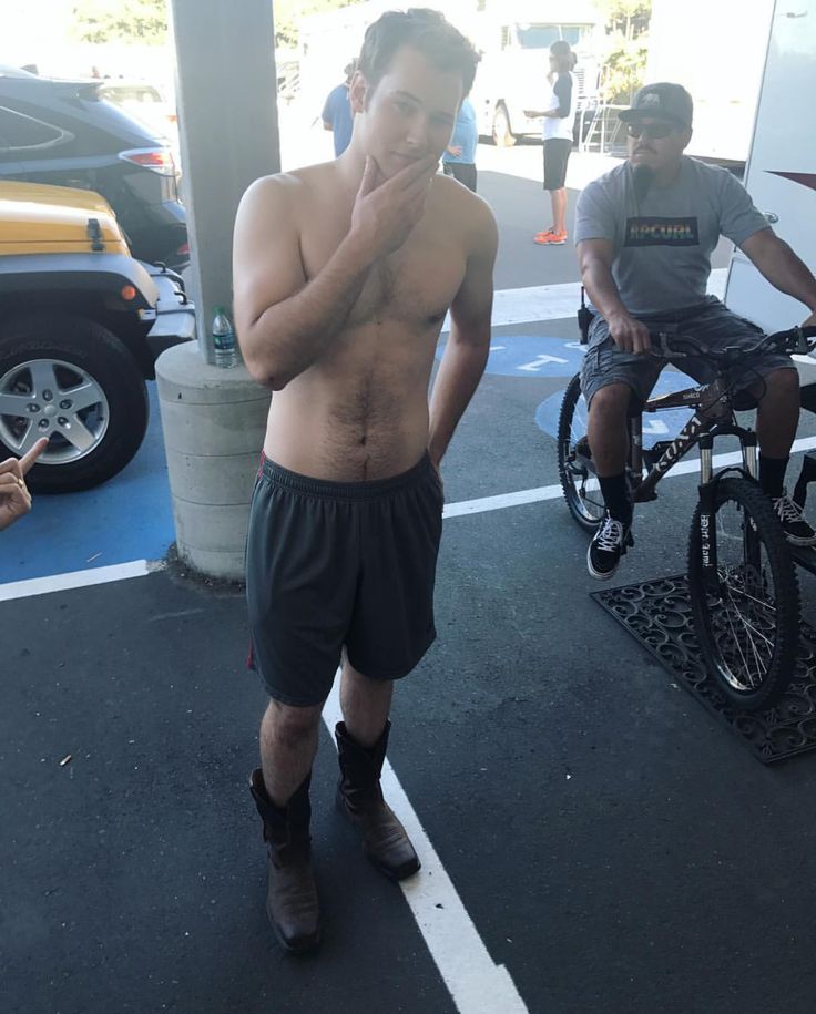 a shirtless man is standing in the parking lot talking on his cell phone while another person rides an electric scooter behind him