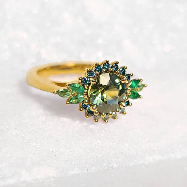 Make a statement with this one-of-a-kind stunner!  It would be ideal as an alternative engagement ring or an everyday cocktail ring.  It is made of eco-friendly 14-karat yellow gold and set with a magnificent Montana sapphire.  The sapphire provides hues of green and yellow and It is accented with blue diamonds and emeralds.  If you are a lover of greenish-blue this 14K Montana Sapphire, Blue Diamond, and Emerald Ring is a match for you! Green Emerald Ring With Halo Round Cut, Green Diamond Topaz Ring, Green Topaz Ring With Diamond Accent, Green Emerald Ring With Halo, Green Diamond Ring With Halo, Green Sapphire Ring With Halo Setting For Promise, Green Diamond Halo Ring In Round Cut, Green Halo Promise Ring, Green Cluster Ring With Halo