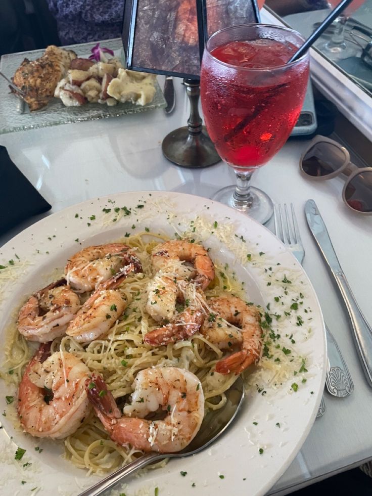 a white plate topped with pasta and shrimp next to a glass of red wine on a table