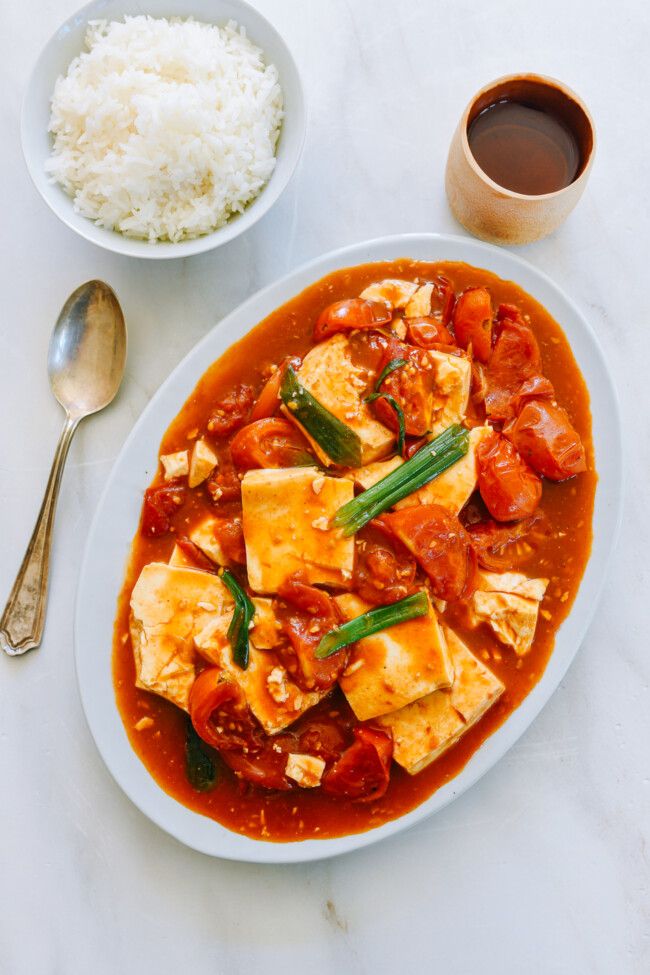 a white plate topped with tofu and rice next to a bowl filled with sauce