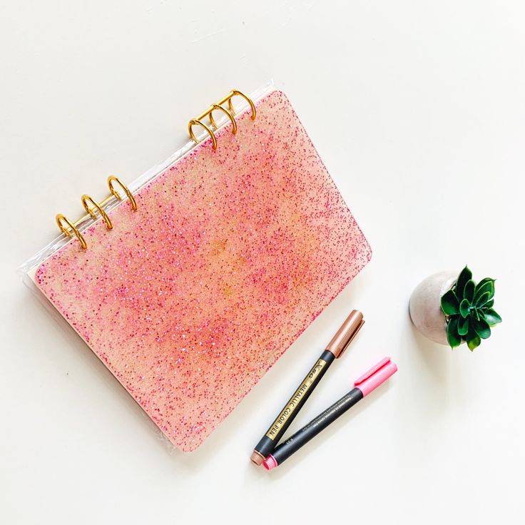 a pink notebook with two pens next to it and a small plant on the side