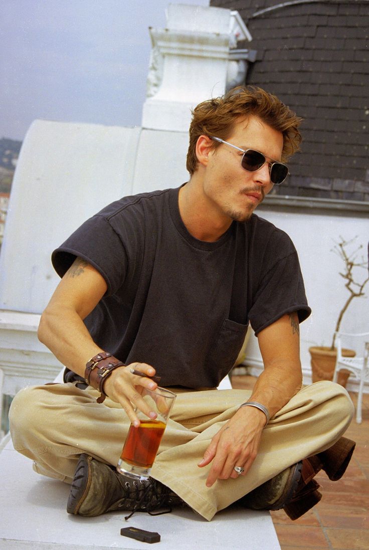 a man sitting on the ground wearing sunglasses and holding a drink in one hand while looking at his cell phone