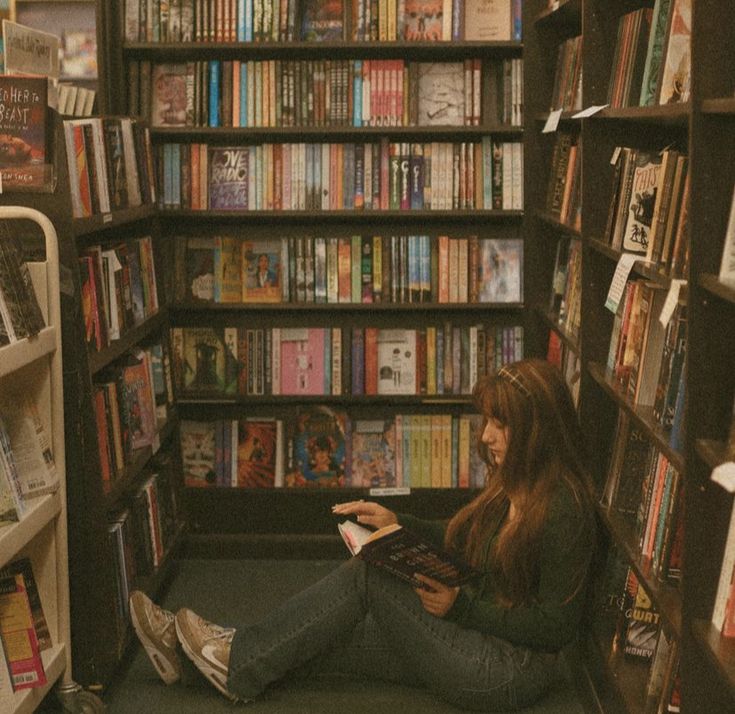 a woman sitting on the floor in front of a bookshelf reading a book