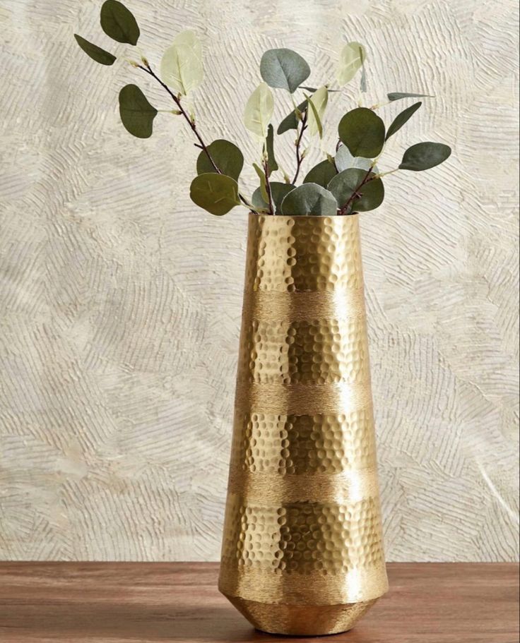 a gold vase filled with flowers and greenery on top of a wooden table next to a wall