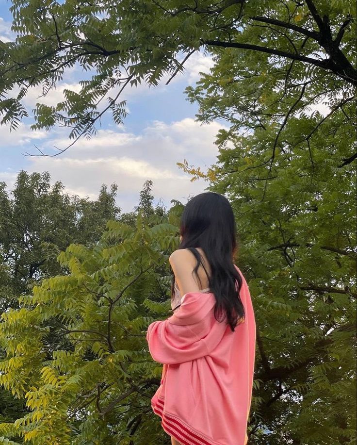 a woman standing in front of trees looking at the sky with her back to the camera