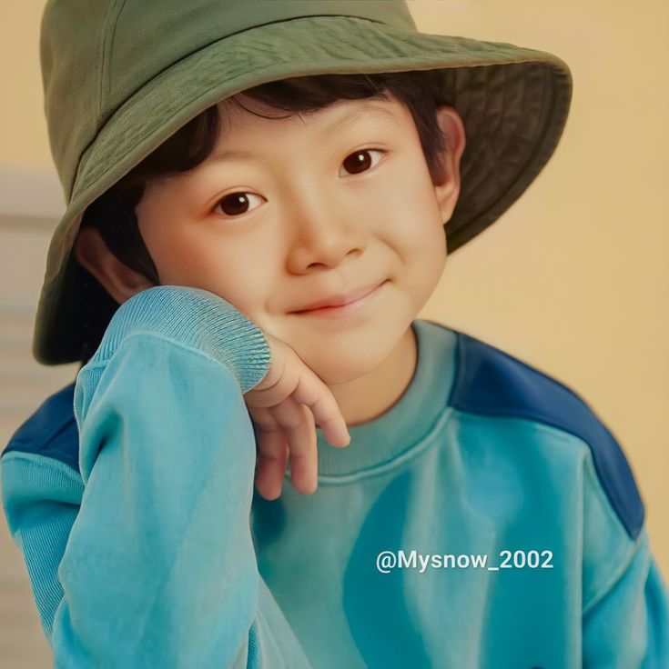 a young boy wearing a green hat and blue shirt with his hand on his chin