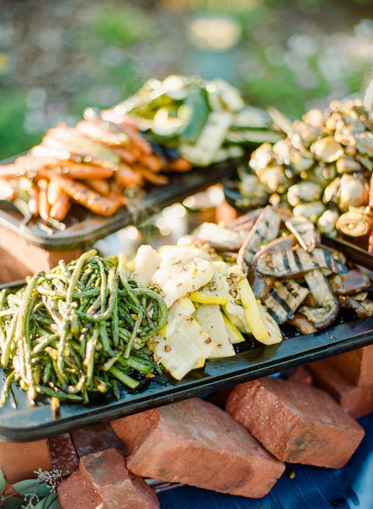 an assortment of meats and vegetables on trays at a buffet table in the park