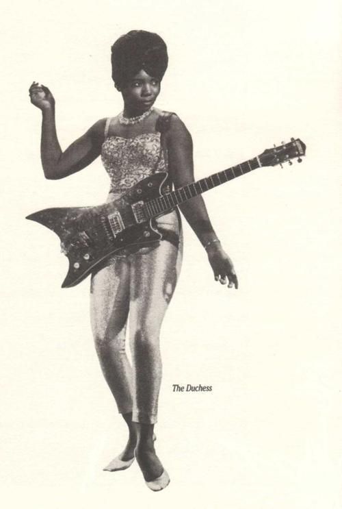 a black and white photo of a woman holding a guitar in her right hand while standing on one leg