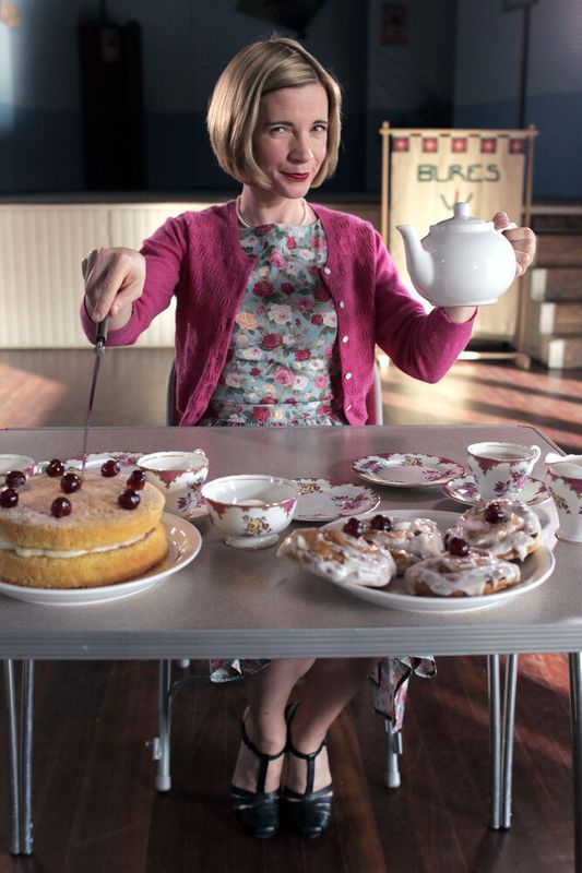 a woman holding a teapot and spoon in front of a table full of desserts