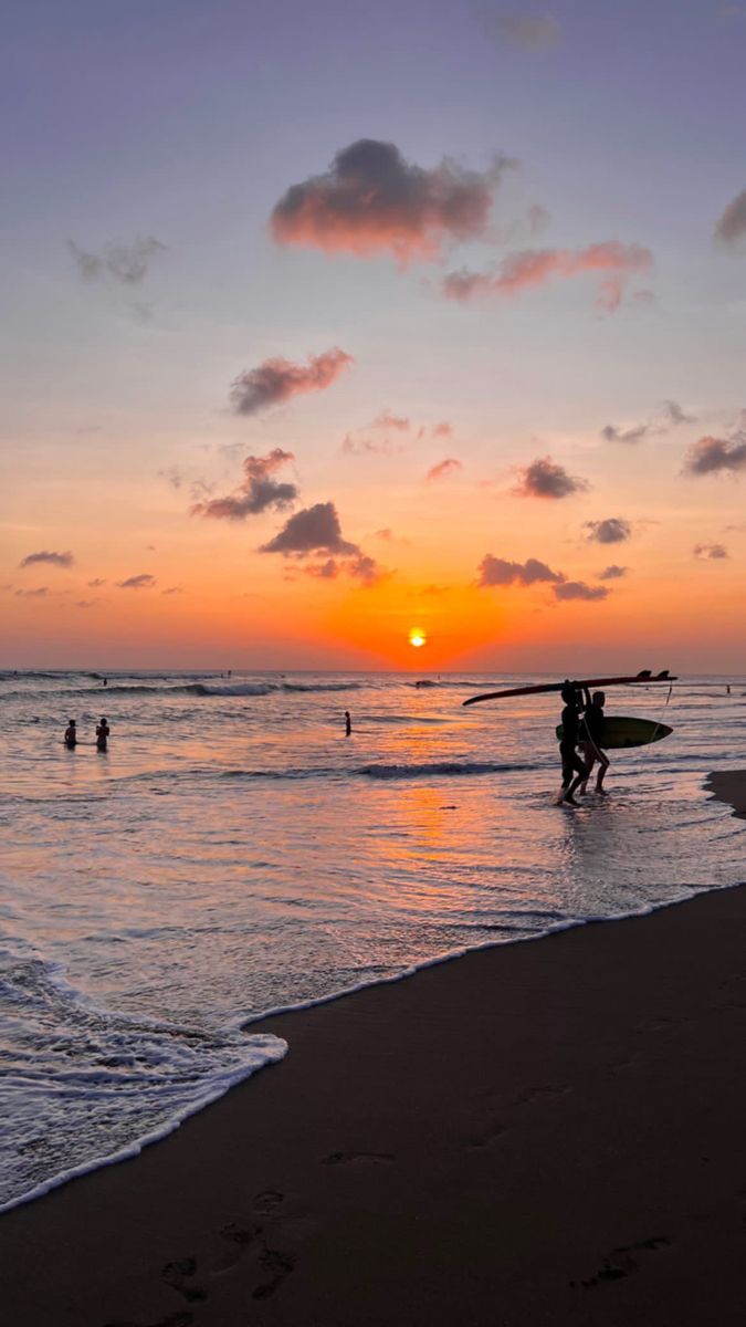 two people carrying surfboards into the ocean at sunset