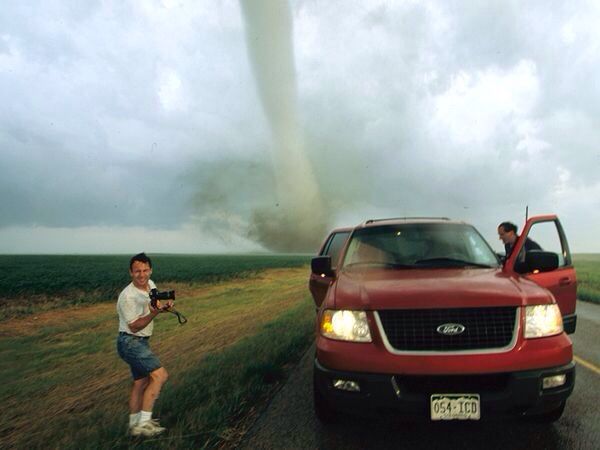 a man standing next to a red truck on the side of a road with a tornado cloud in the background