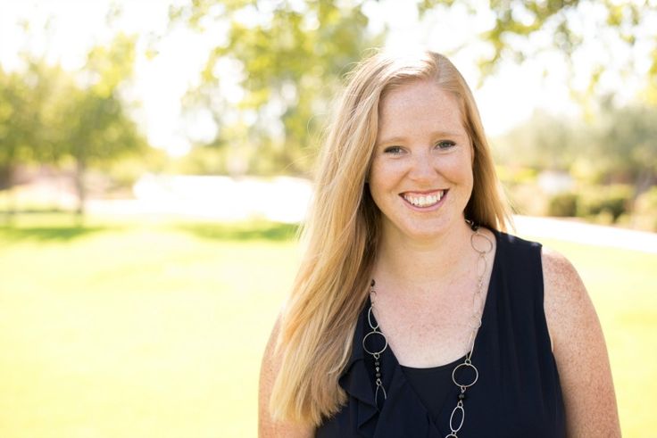 a woman with long blonde hair standing in front of a tree and grass field smiling at the camera