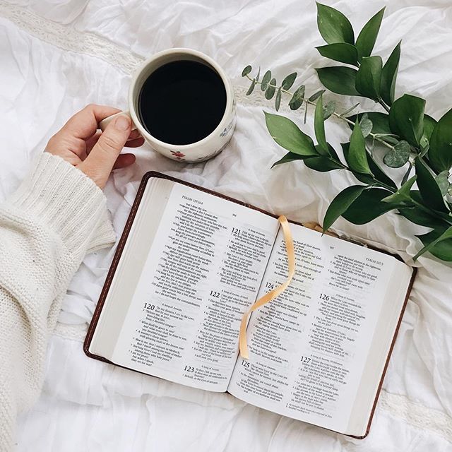 an open book and cup of coffee on a bed with white sheets, greenery