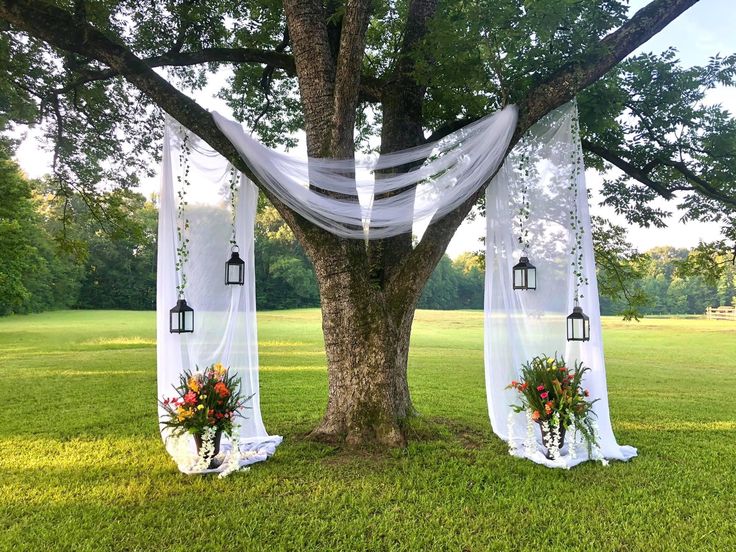 an outdoor ceremony setup with white drapes, lanterns and flowers on the grass under a tree